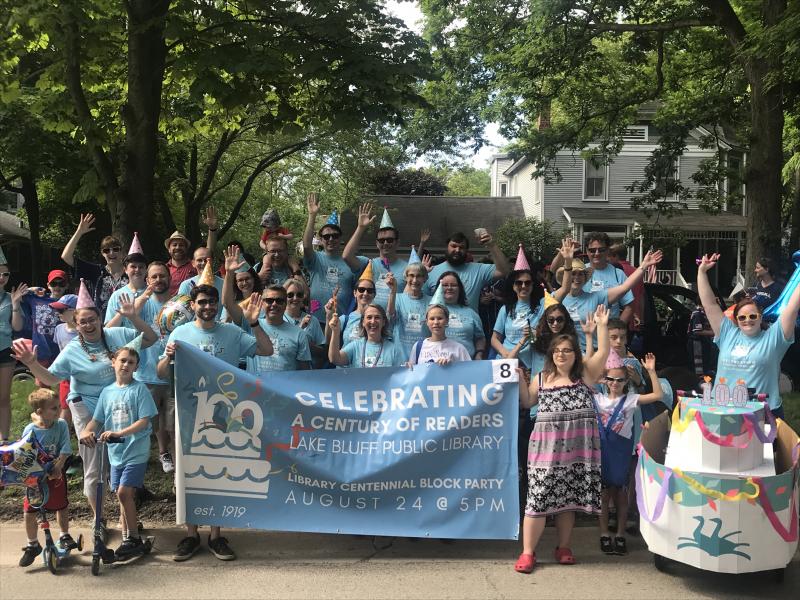 A group of people in blue shirts and party hats standing in front of a sign reading Celebrating a Century of Readers Lake Bluff Public Library Centennial Block Party August 24 at 5pm.