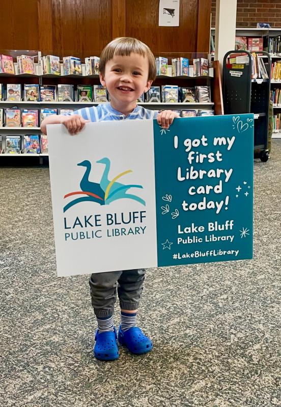 Child standing in the Youth Services Department holding sign "I got my first library card today!"