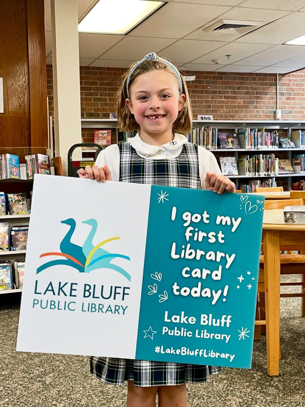 Child standing in the Youth Services Department holding sign "I got my first library card today!"