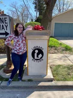 A woman standing in front of a drive up Library book drop with a red bow on it.