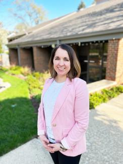 A woman in a pink blazer standing in front of a library and smiling.