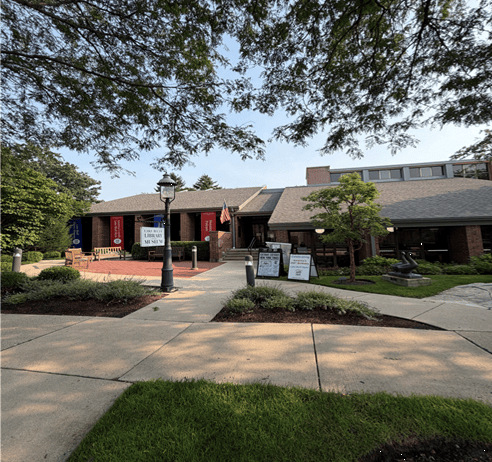 A brick building with red and blue banners, signs out front, and trees and bushes along a sidewalk under a sunny sky.