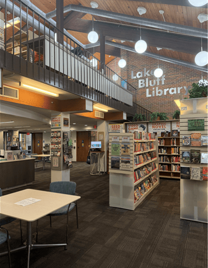 Interior of Lake Bluff Library with bookshelves, tables, chairs, and a modern, airy design. Large windows and globe lights brighten the space, with Lake Bluff Library on the brick wall in the background.