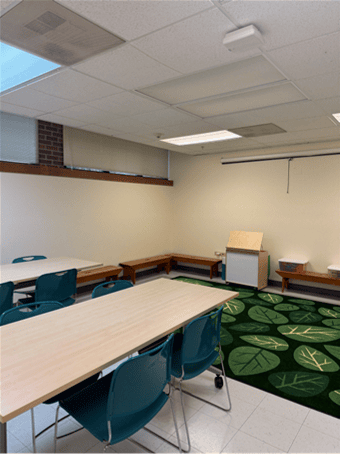 A classroom with light wood tables, teal chairs, benches along the walls, and a green rug with leaf patterns. Theres a small white podium and natural light from ceiling windows.