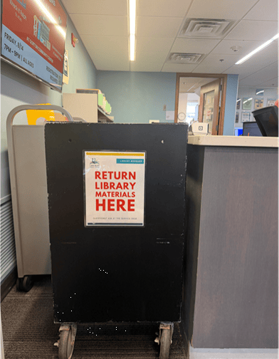 A black cart with a sign reading Return Library Materials Here stands beside a counter in a library. A book cart and library signage are visible in the background.