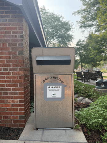 A metal library return box stands by a brick wall outside, with a sign reading NO DONATIONS and greenery nearby. Benches and trees are visible in the background.
