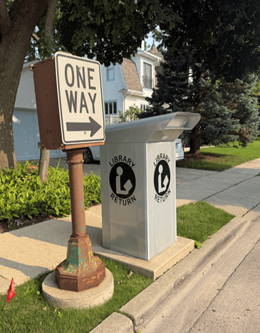 A one-way street sign stands beside a sidewalk library return drop box in a suburban neighborhood with houses, trees, and grass visible.