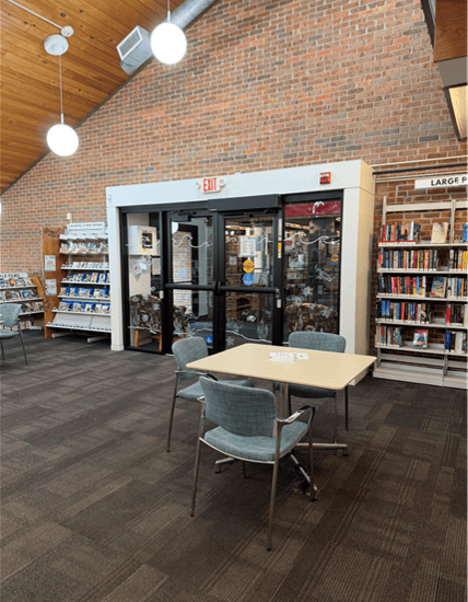 A library interior with bookshelves, a table with four chairs, and glass double doors. The ceiling is wooden with hanging lights. A sign on a shelf reads LARGE..