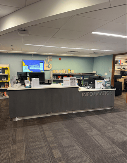 Information desk in a library with computer monitors, brochures, and signs. Bookshelves and a display rack are visible behind the desk. The area is well-lit with gray carpeting and a blue accent wall.