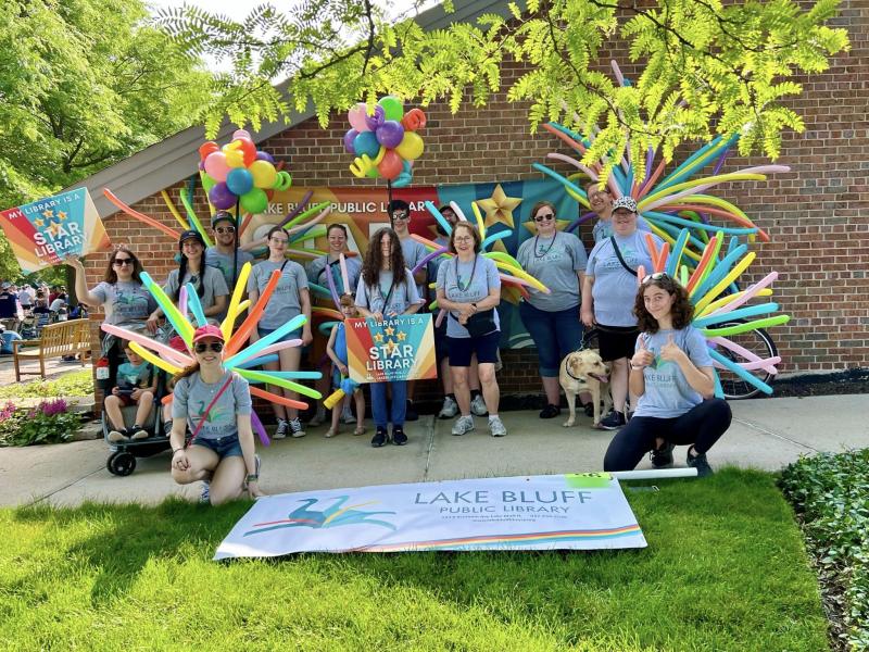 A group of people with colorful balloon decorations pose in front of a Lake Bluff Public Library banner outside a brick building, celebrating as a Star Library. Some hold a sign and balloons; a dog is present.