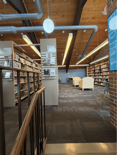 A modern library interior with bookshelves, a wooden ceiling, exposed ducts, posters, and a seating area with cream-colored chairs in the background.
