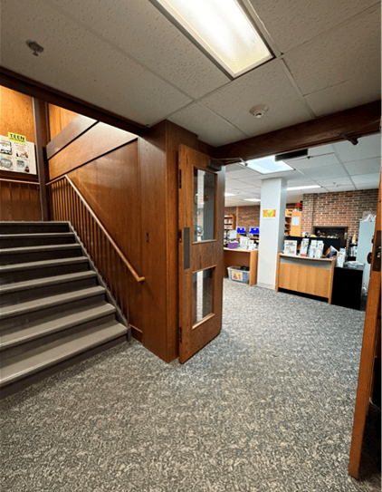 A hallway with carpeted floors, a wooden staircase on the left, and an open door leading to an office area with desks, shelves, and various supplies visible in the background.