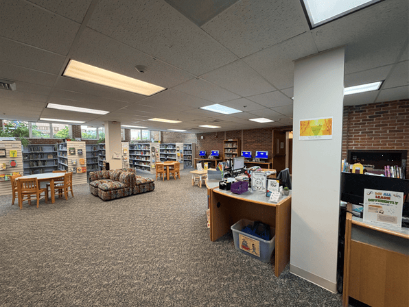 A library interior with bookshelves, computer stations, a patterned couch, and wooden tables with chairs. Large windows and overhead lights brighten the open, carpeted space.