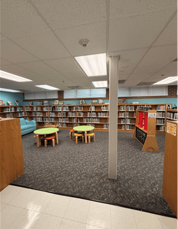 A library childrens area with low bookshelves, round tables, small stools, a blue couch, and a carpeted floor. Bright lights and books line the walls.