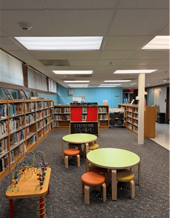 A childrens library area with low bookshelves, small round tables with orange stools, a bead maze toy, and a sign that says Storytime Corner. The space is well-lit with overhead lights.