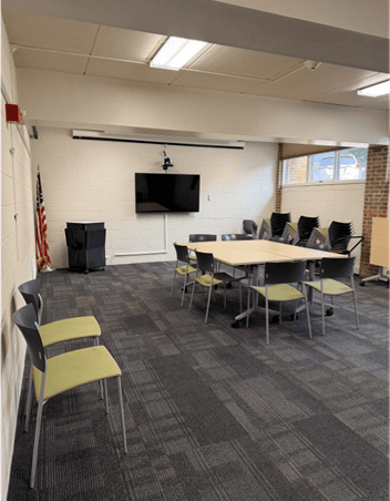 A meeting room with tables arranged in a square, chairs around and stacked, a TV on the wall, an American flag in the corner, and windows letting in natural light.
