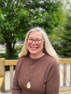 A smiling woman with long gray hair and glasses sits on a wooden bench outdoors, wearing a brown sweater and a necklace. Green trees and plants are blurred in the background.