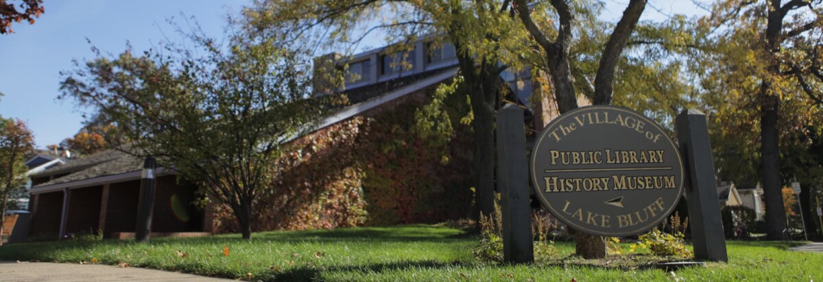 A round sign for The Village of Lake Bluff Public Library and History Museum stands on a grassy lawn in front of a brick building with trees in the background.