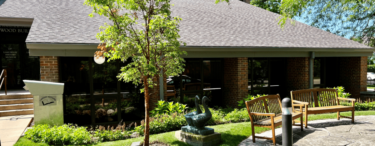 Two wooden benches and a bird sculpture sit on a stone patio outside a brick building surrounded by green plants and trees, with sunlight casting shadows on the ground.