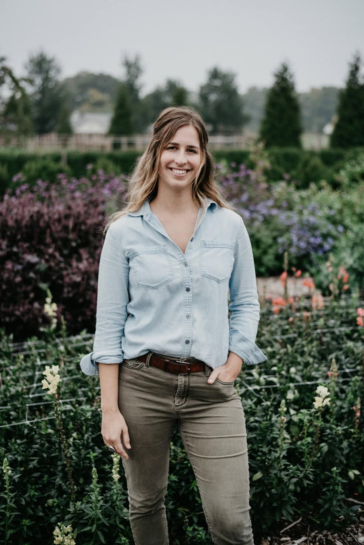 A woman with long hair, wearing a light blue shirt and khaki pants, stands outdoors in a garden with colorful flowers and green plants, smiling at the camera.