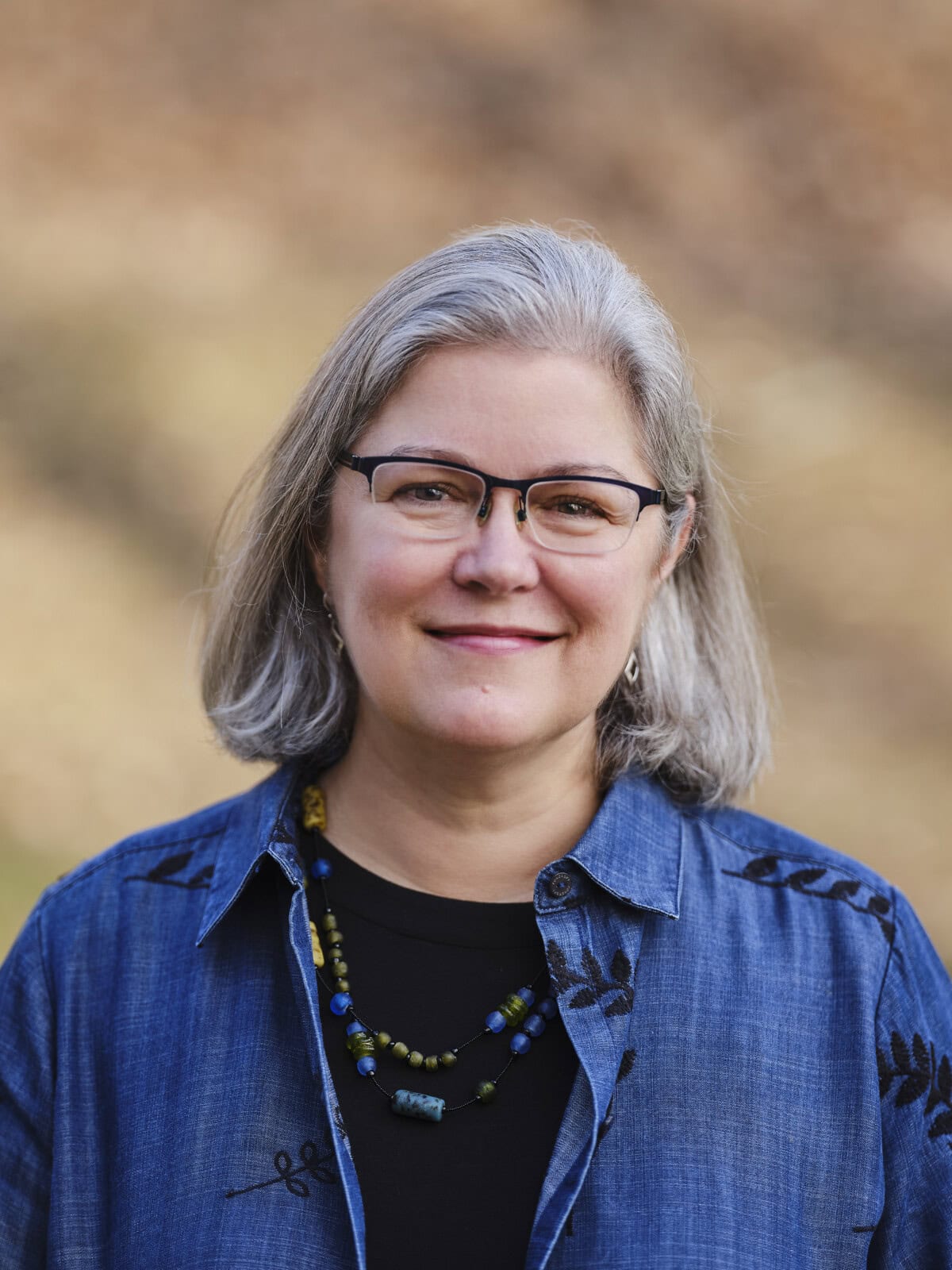 A woman with gray hair and glasses smiles at the camera. She is wearing a blue shirt, a black top, and a beaded necklace. The background is out of focus with brown and tan tones.