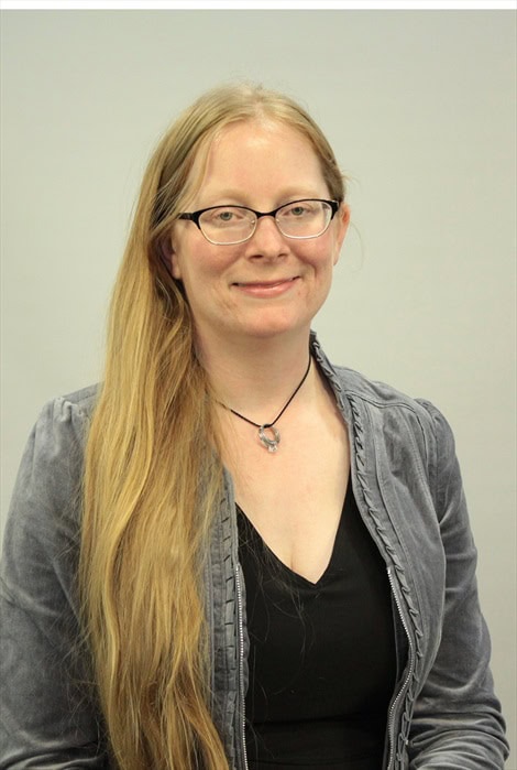A woman with long blonde hair and glasses, wearing a grey jacket over a black top and a necklace, smiles slightly against a plain light gray background.