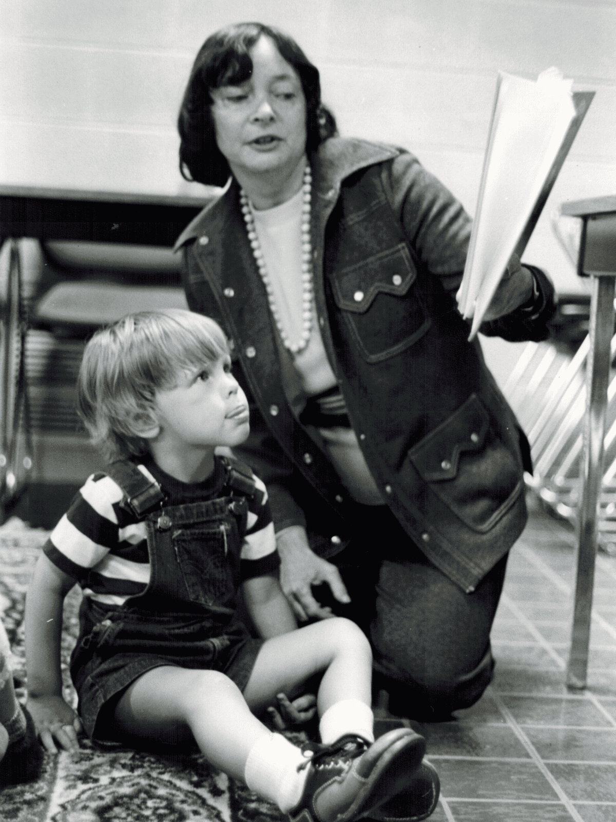 A woman kneels beside a young boy sitting on the floor. She is holding open papers while the boy looks up at her. They appear to be in a classroom with chairs stacked in the background.