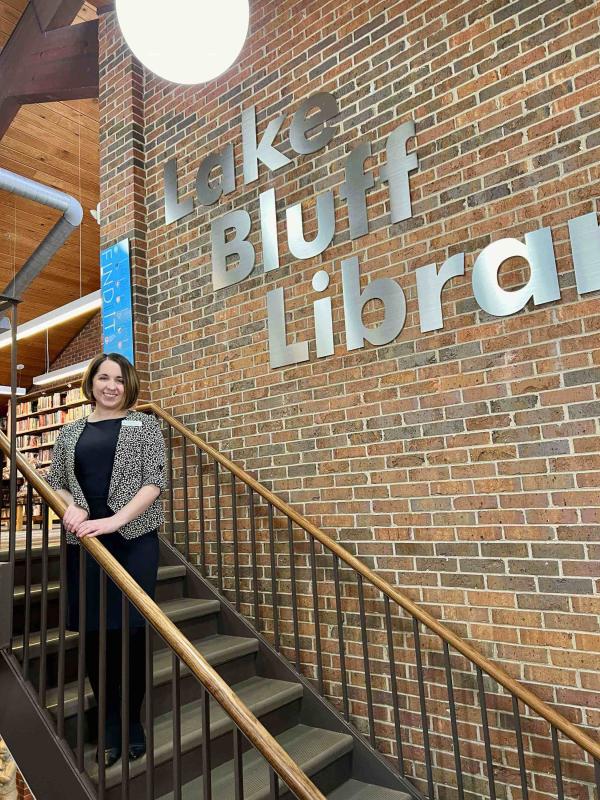 A woman stands smiling on a staircase inside a library, next to a large wall sign that reads Lake Bluff Library. Shelves of books are visible in the background.